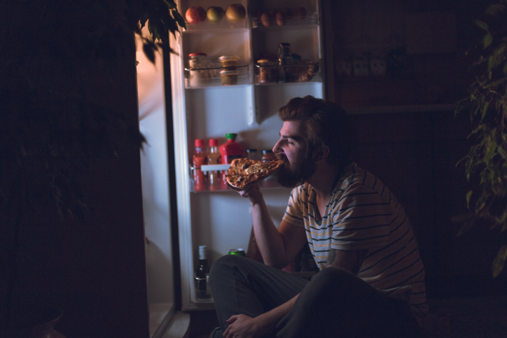 Un homme mange une pizza pendant la nuit, assis devant le réfrigérateur.